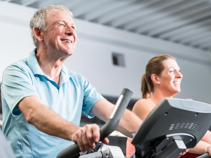 Älterer Mann und eine Frau trainieren lächelnd auf Ellipsentrainern in einem Fitnessstudio.
