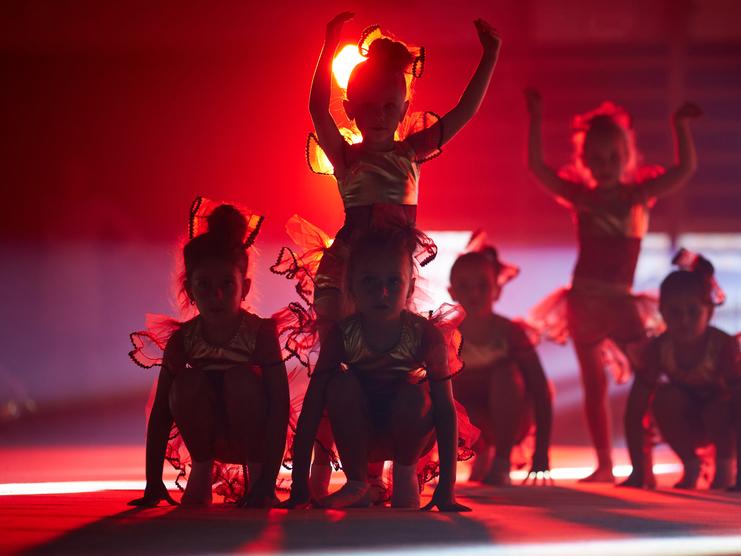 Sechs Kinder in goldenen Kostümen mit roten Rüschen posieren in rotem Licht auf der Bühne.
