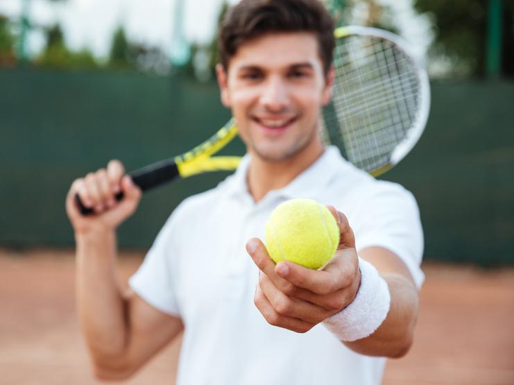 Lächelnder Mann in Sportkleidung hält einen Tennisball in der Hand, mit einem Tennisschläger über der Schulter.