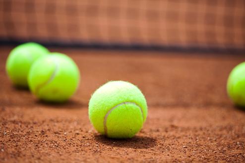 Vier grüne Tennisbälle liegen auf einem roten Sandplatz, mit einem Netz im Hintergrund.