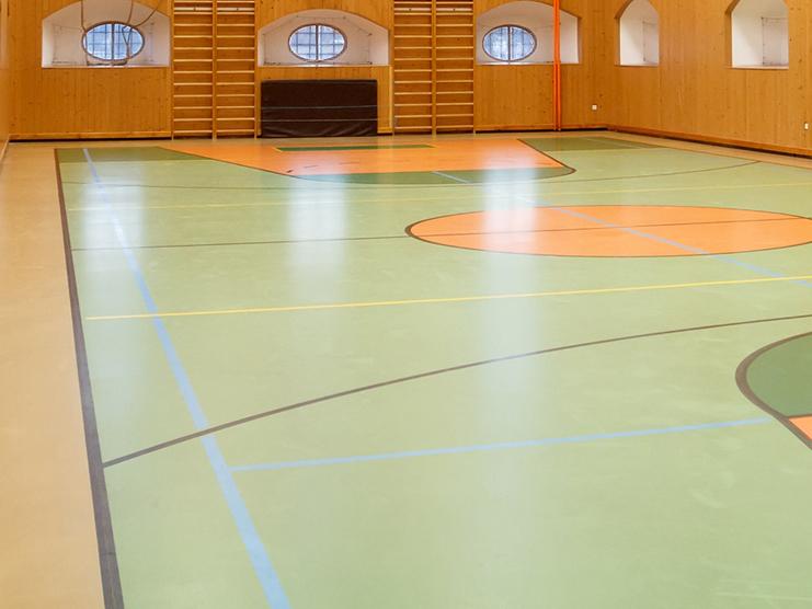Helle Sporthalle mit Holzverkleidung, Basketballkorb und farblich markierten Spielfeldern auf dem Boden.